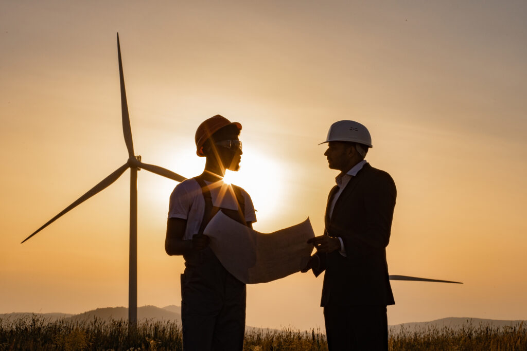 Dwóch wielorasowych pracowników przemysłowych badających plany turbin wiatrowych z niesamowitym zachodem słońca w tle. Obszar wiejski z ekologiczną farmą produkcji czystej zielonej energii.
