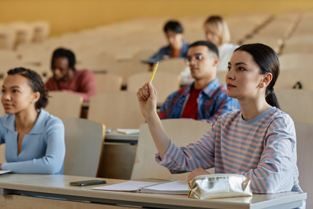 Studentka podnosi rękę, aby zadać pytanie, siedząc przy biurku z kolegami i słuchając wykładowcy